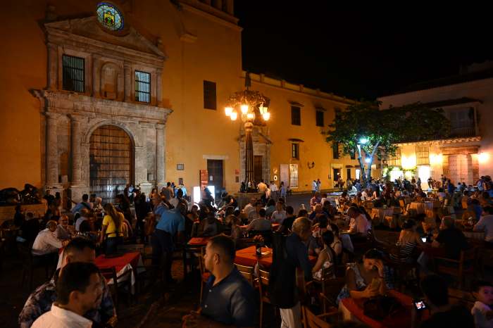 Central Square Cartagena Colombia at night, with lots of outdoor seating, lots of people seated in front of the old colonial architecture, elaborate city lights, and with an overall super charming night atmorphere