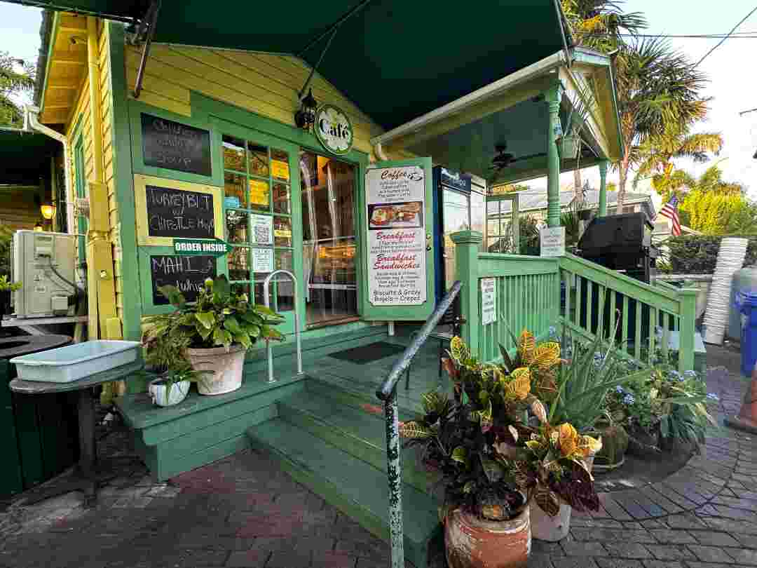 Charming cafe in an old wooden house painted in yellow and green in Key West. The house is surrounded by plants and flowers, and has old fashioned rustic signs on the porch up the two three steps from the street. 