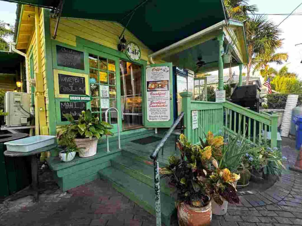 An incredibly cute breakfast and coffee shop in a green and yellow wooden old house with crooked stairs, lots of flowers and plants outside, and an old cafe shop above the door. 