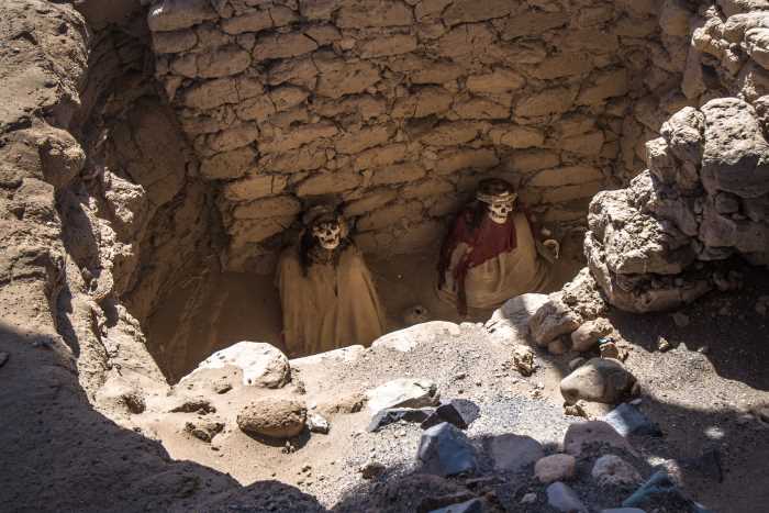 The mummies in Chauchilla Cemetery sitting in an open grave looking up, with white faces, brown cloaks, and head bands in the middle of a rocky grave. 