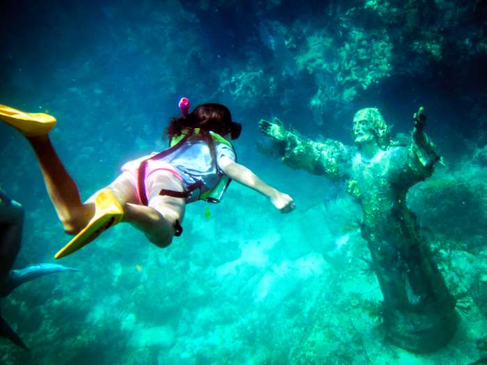 The greenish clear waters outside Key Largo is perfect for snorkeling, here there is an underwater statue of a man that resembles Jesus standing on the bottom, and a snorkeler in colorful equipment is on her way down to it. 
