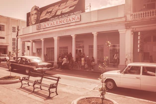 An old photo from Cienfuegos street ambiance in the 50s, with old fashioned cars in the street and people gathered outside a venue with a large photo of Che Guevara over the entrance