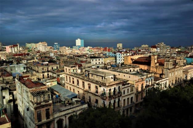 Rooftop view of Central Havana from Iberostar Parque Central terrace, lots of rugged houses and structures towards the Malecon and the sea under a darkening sky while the sun is still shining on the buildings