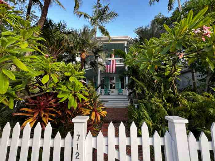 The white wooden gate to a charming wooden house in Key West, surrounded by greenery, and the American flag on the porch under the blue sky
