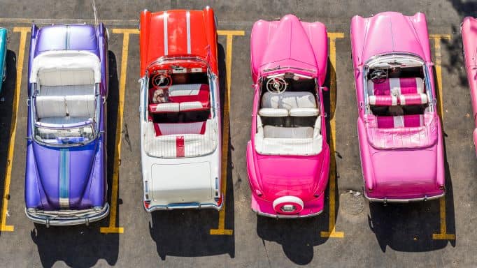 A row of classic American cars, convertibles in bright colors, seen from above. One purple, one red, and two bright pink. 