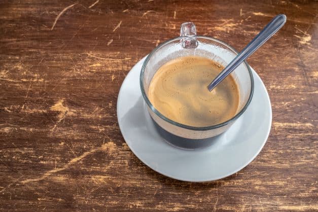 Tiny glass cup of strong sweet coffee on a white plate, with light foam on top, and a spoon to dessolve the sugar