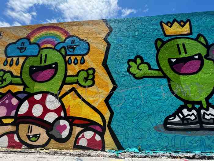 Close up photo of Street art in Wynwood, colorful cartoon like cural with green creatures with a big smile on a yellow and blue background respectively, under the blue sky. 