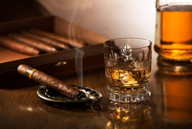 A lit cigar lying in an ashtrey, next to a decorative glass with golden rum inside, on a dark shiny wooden table