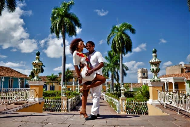 A salsa dancing couple dressed in white in a park in Cuba, on a bright sunny summer day with greenery and palm trees under a clear blue sky. 