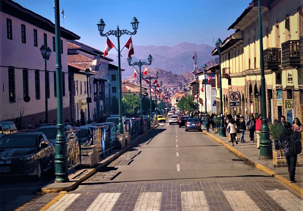 The main street of Cusco Peru, a wide street with cars and pedestrians, bathed in sunlight. You can see the mountains in a distance. 