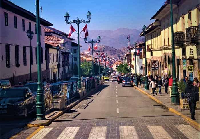 The main street in Cusco on a sunny but cold day, with old stone houses on both sides, flags along the road, and mountains in the distance far away