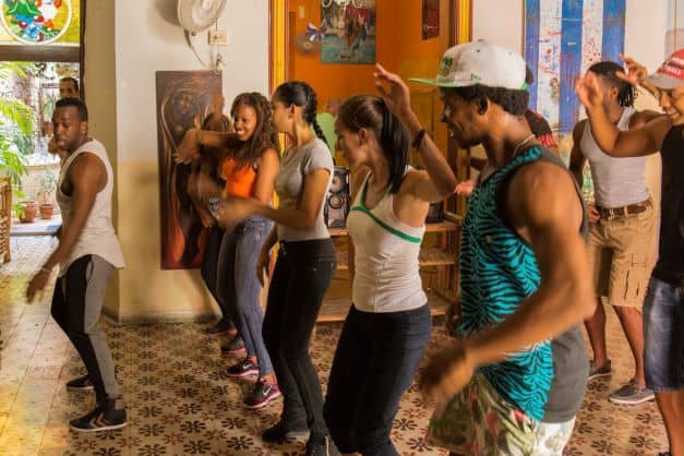 Dance class at Casona del Son in Old Havana, Cuba. A group with a teacher is dancing in a beautiful room, with decorative tile floors, art on the walls, and colored glass details on the doorways. Everyone smiling and having fun. 