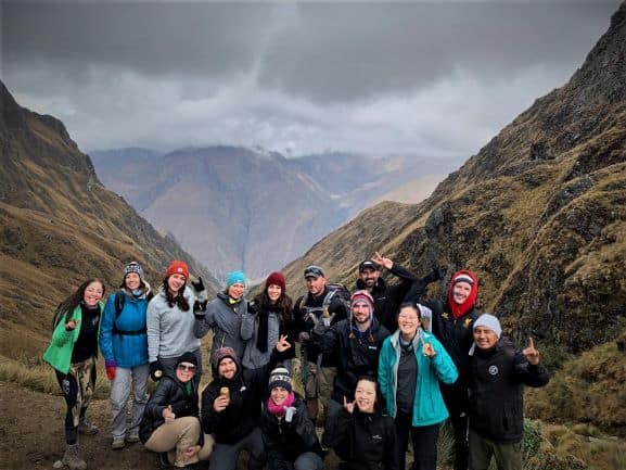 Everyone at the highest peak altitude in the middle of the Dead Womans Pass. There are still hillsides higher around us, and there are infinite hilly mountains in the distance in a hazy air. 