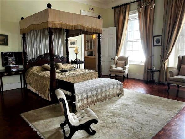 A very stylish bedroom at Devon House, with a lush white carpet, king bed, elegant furniture, floor to ceiling curtains, and a dark elegant wooden floor. 