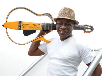 The "father of Kizomba" Edward Paim, smiling at the camera wearing a white t-shirt, a hat, and his buitar across his right shoulder. 