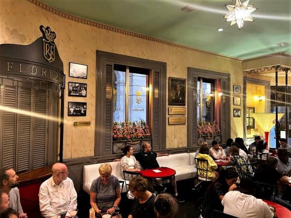 The seating area in Floridita Bar in Havana, with lots of people seated in the white sofa by the red small tables, with charming classic colonial windows where you can peek out to the street outside