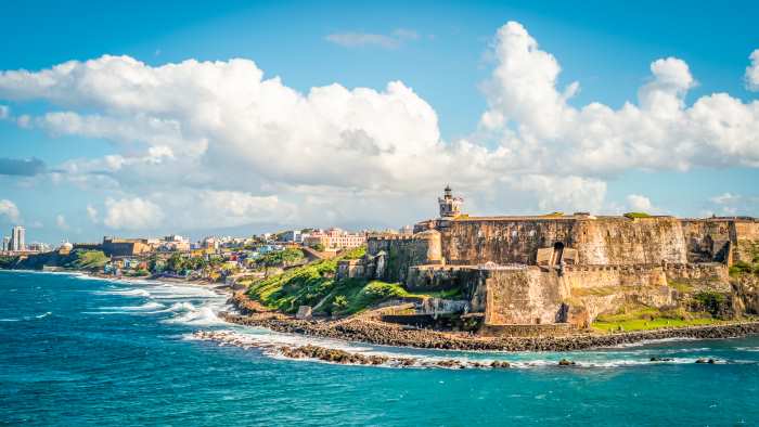 The fortress and lighthouse at the tip of San Juan in Puerto Rico, sitting high on a cliff guarding the entrance to the city. The photo is taken on a hot summer day, with blue skies, and blue waters below the fortress. 