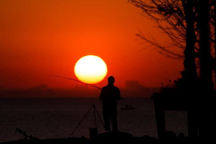 A man fishing with a rod in front of a glowing red sunset with a huge sun in hte background, surrounded by a glowing red sky