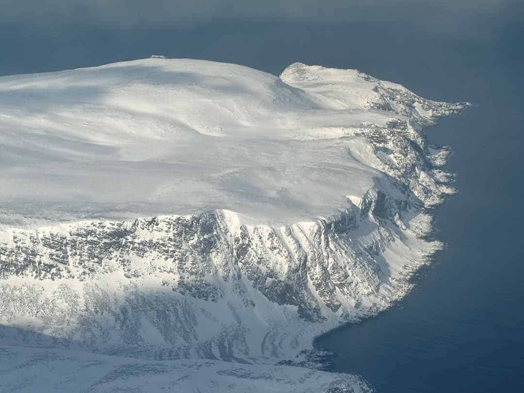 Flying over the white snow covered North Cape area in Finnmark, Norway. The rugged mountain is bright white against the deep blue almost black sea, bathed in sunlight. 