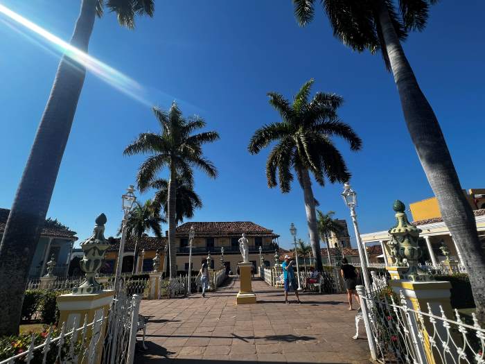 The Plaza Mayor Park on a bright sunny day, with palm trees, white iron fences, statues, and people strolling the park. 