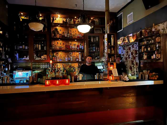The bar at the Freehand Miami, with warm dimmed lighting, a dark wooden bar with a white stone counter, and a myriad of bottles behind the bartender who is smiling inside the bar