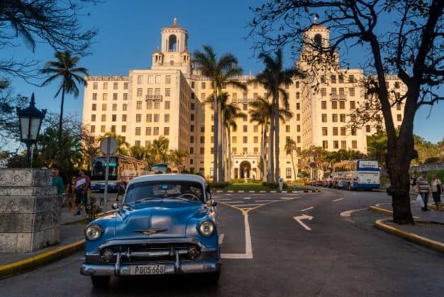 The impressive facade and entrance to Hotel Nacional de Cuba, bathed in sunlight, and a blue classic American car is driving out the entrance. 