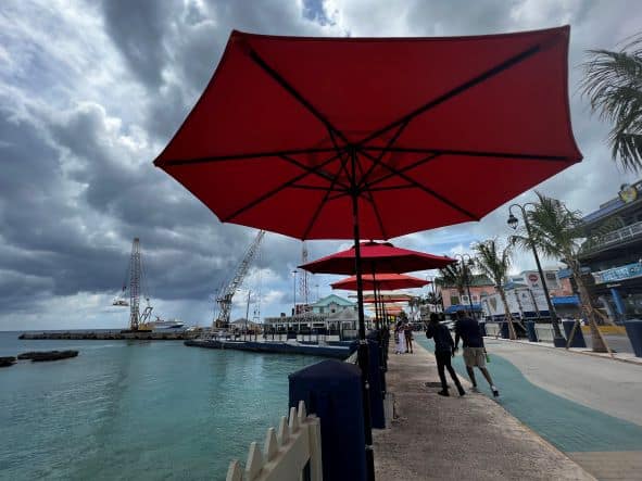 Red parasols lined along the boardwalk in George Town on a cloudy day, people walking along the sea amidst houses and palm trees. 