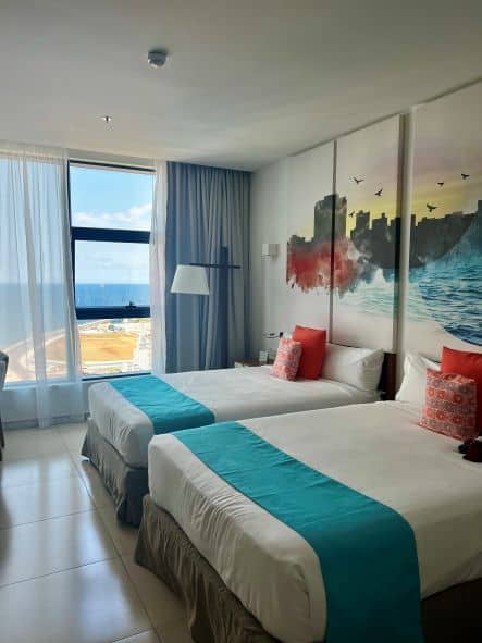 Elegant airy rooms at the Grand Aston, with two queen beds with white linen, blue bed covers and red pillows. You can see the park by the Malecon outside the window. 