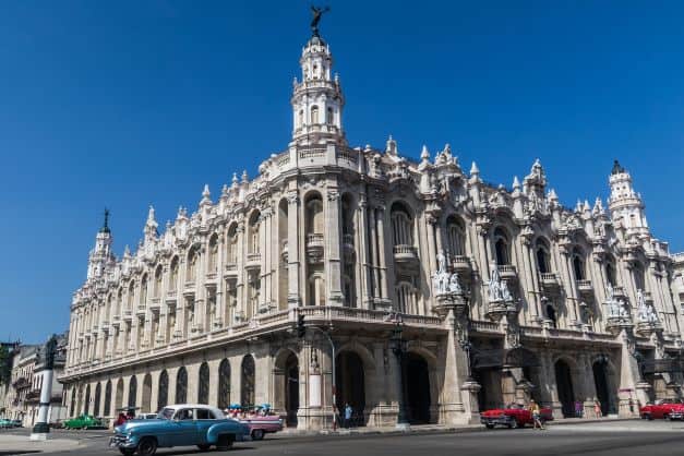 The beautiful white building that houses the Grand Theater of Havana. With speers and columns and ornate art details, under a deep blue summer sky. 