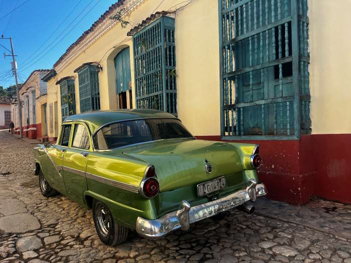 A green metallic classic american car parked on a cobblestoned street in Trinidad, next to colonial houses painted in light yellow and red, with green wooden window shields