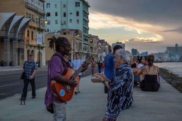 The Malecon boardwalk in Havana around sunset, with a guy playing the guitar in front of the colonial houses, lots of people seated on the brick wall, and the sun lights up the clouds in the distance. 