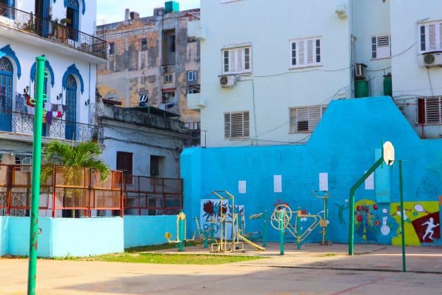 A mix of playground and basketball court in Old Havana, with bright light blue walls and different play stations amidst run down and still beautiful houses. 