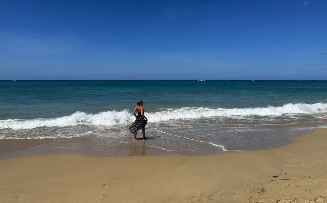 Me, a solo female traveler, walking in the soft surf alone on the golden sandy beach in San Juan Puerto Rico on a bright summer day with blue seas and blue sky.