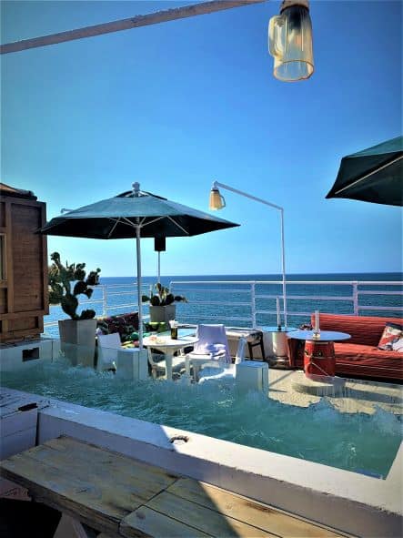 Malecon 663 Rooftop in Havana on a bright summer day with the blue sea in the distance, and blue skies. The terrace has seating areas, a red sofa, circular tables, and a glass jacuzzi. 