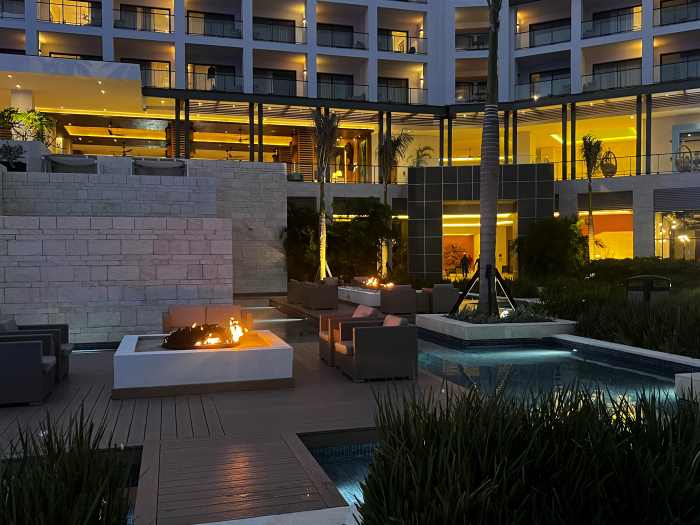 One of the zones around the pool area at night with warm lighting, blue lights from the smaller pools, seating areas and little fires, surronunded by wood flooring and white stone structures. 