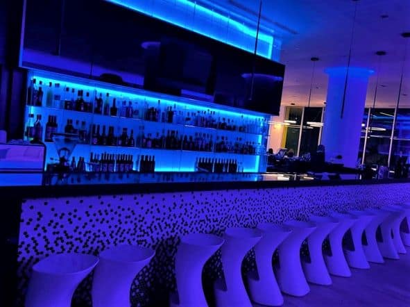 The blue light bar at the Iberostar Grand Packard, which is on the same floor as the pool and a restaurant. The bar has white stools lined for guests to sit, and the bar shelves behind the bar are also lit in blue. 