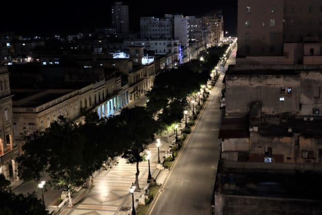 The venerable Prado Avenue at night, separating Old and Central Havana. The avenue is lit with elegant lights, and rows of big trees shelter the pedestrian walkway with roads on each side. 