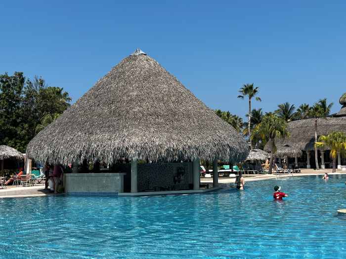 A large blue pool in a Varadero resort with a charming pool bar with straw roofs amidst holidaymakers enjoying the warm sunny summer day