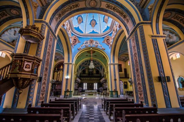 The beautiful interior of Santiago de Cuba cathedral, with high curved ceilings, colorful art in yellow, red and blue, intricate patterns on the walls and ceiling, and lots of light coming in. 