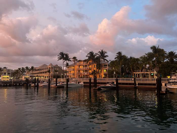 Sunset over Key West Marina, with the dark water in the foreground, and the jetty with all the restaurants and buildings surrounded by palm trees in a golden light in the background, under a darkening sky