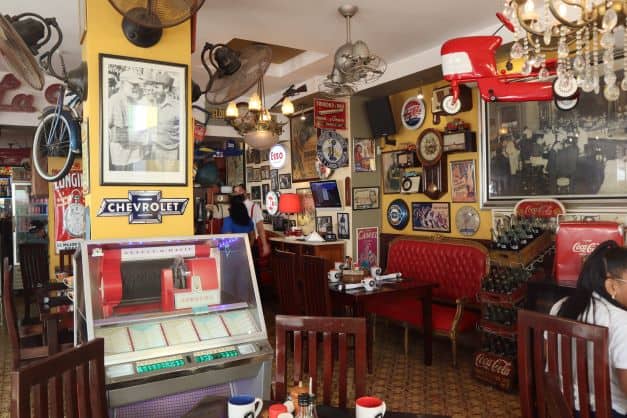 La Vitrola interior which is a mix of eclectic details and ornaments on the wall, and old music box, car signs, photos, an old red sofa, and seating areas with wooden furniture. 