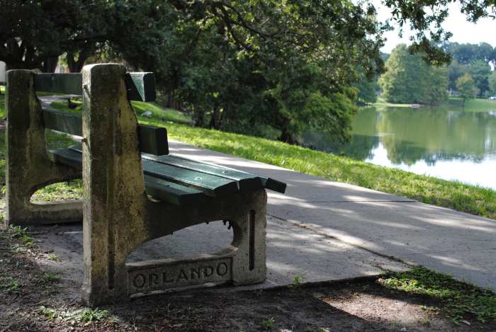 A cute park bench of stone and green wood by the calm Lake Davis in Orlando on a sunny summer day, with green trees and bushes lining the lake shores