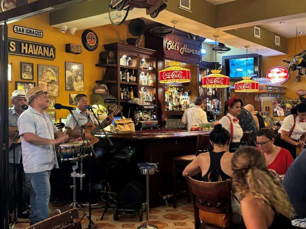 A vibrant bar in Little Havana with live music, lots of guests, and a busy bar with colorful yellow walls, brown bar, and details in red and yellow. 