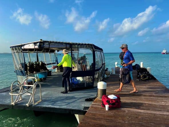 Heading out for a day of scuba diving in Grand Cayman, everyone prepping for the day carrying gear onto the small dive boat that is docked on a small wooden jetty. It is a bright summer day, blue seas and blue skies! 