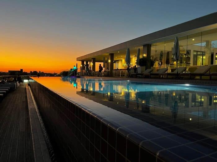 Manzana Kempinski rooftop pool in Havana just around sunset. The pool is lit from below, the water is blue, the elegant restaurant is in the background, and the sky is glowing orange from the sunset.