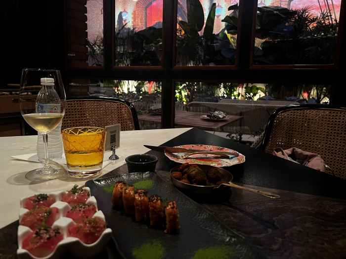 An elegant sushi restaurant with warm intimate lighting in Zona Colonial, Santo Domingo. On the table is a variety of sushi dishes and a glass of white wine, and in the back plants in the indoor atrium. 