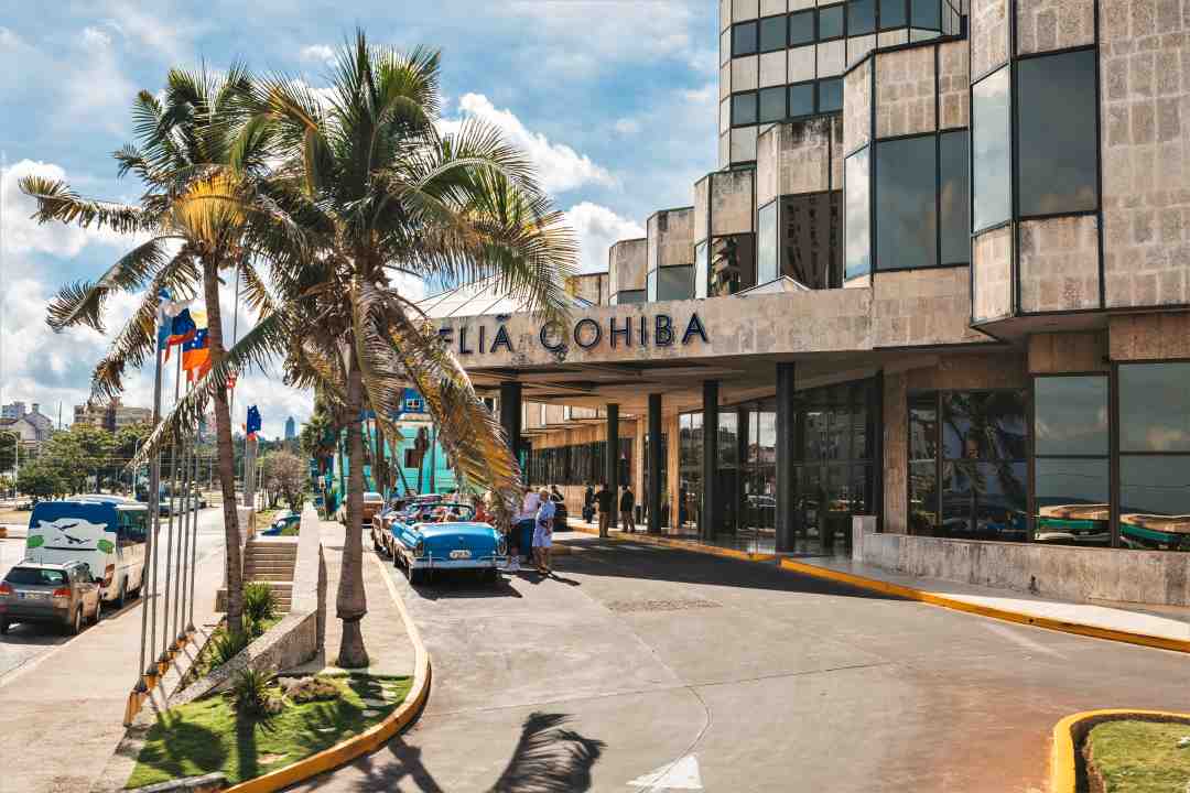 The drive-in entrance to the Melia Cohiba in Havana on a bright summer day, with a large reception area, classic cars parked outside, and a row of flags by the palm trees. 
