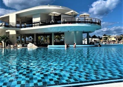 Pool at a luxury resort in Varadero Cuba, blue pool water where you see the chess patterend bottom shining through. On the far side are people relaxing in the pool bar, under a blue sky on a sunny day. 