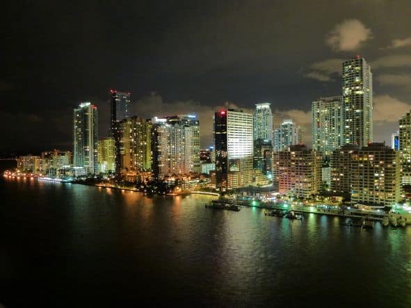 The Miami skyline at night, the sea and sky is dark, and the skyline is beautifully lit with thousands of lights from the buildings, streets and cars. 