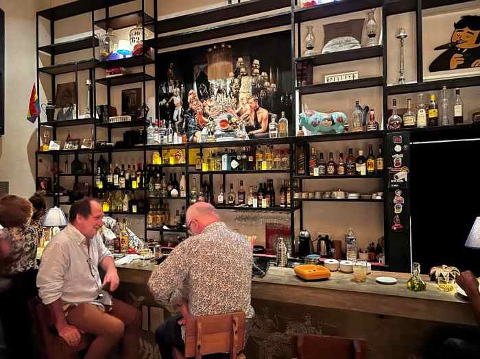 The bar area in Paladar Mischifu, a wooden bar, and behind it all the bottles and drinks are on black shelves, some with lighting. Along the bar are seated customers enjoying themselves. 